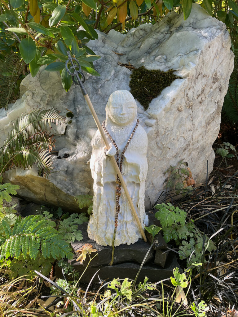 A statue of a Jizō, a protector bodhisattva. The statue is a white figure whose face is at peace eyes closed contentedly and smiling, while draped in mala prayer beads and holding a staff serenely. Even though his body is crumbling, dissintigrating, this does not impact his sense of peace and wellbeing. 